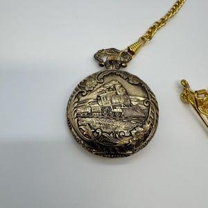 May include: A gold-toned pocket watch with a detailed train scene. The watch has a decorative chain and clasp. The watch face features an embossed image of a steam train, set against a background of floral and scrollwork.