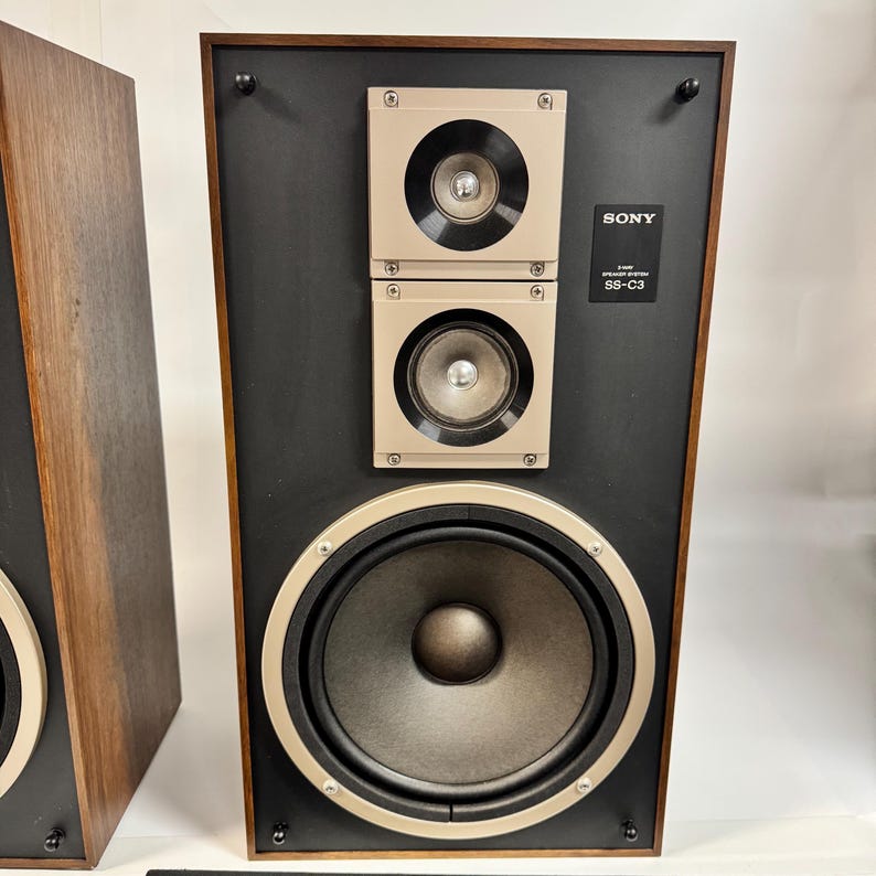May include: Vintage Sony SS-C3 speaker system. The speaker features a black face with a large central speaker and two smaller speakers above. The speakers are framed in a gold-colored metal and set in a wooden cabinet.