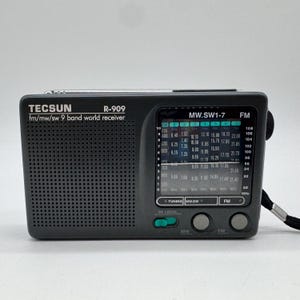 以下が含まれることがあります： ダークグレーのTecsun R-909 FM/MW/SW 9バンドワールドレシーバーラジオ。左側にスピーカーグリル、白い文字の周波数表示パネル、チューニングダイヤルがあり、右側には黒いリストストラップが取り付けられています。