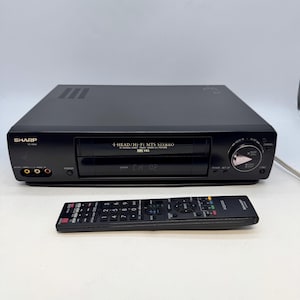 May include: A black Sharp 4-Head Hi-Fi MTS Stereo VHS HQ video cassette recorder with a front-loading tape slot, power button, and input jacks. It includes a matching black Sharp Aquos remote control with numerous buttons for playback and menu navigation.