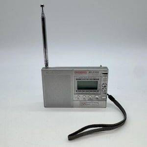 May include: A silver portable radio with a long telescopic antenna, a digital display, and a speaker grille. The device features buttons for power, light, sleep, and memory, with the text KK-C300 PLL SYNTHESIZED FM STEREO/MW/SW RECEIVER.