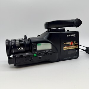 Könnte beinhalten: Ein schwarzer Sharp Camcorder mit 8-fach-Zoomobjektiv, CCD-Sensor und einem oben montierten Sucher. Das Seitenpanel zeigt den Text Power 8x Zoom und One Touch Full Auto sowie verschiedene Bedientasten und ein kleines digitales Display.