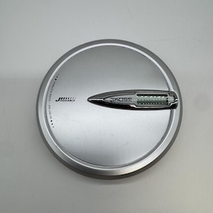 Może przedstawiać: Okrągły, srebrny przenośny odtwarzacz CD z metalicznym wykończeniem. Urządzenie posiada nadrukowaną na czarno nazwę marki Koss oraz tekst Joggable i 60 SEC ASP / CD-RW PLAYBACK. Pośrodku znajduje się poziomy panel wyświetlacza.