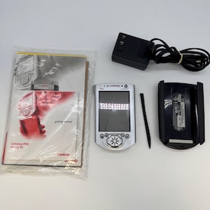 May include: A silver Compaq iPAQ Pocket PC with a stylus, a black power adapter with a cord, a black cradle, and a printed getting started manual in a plastic sleeve. The device features a screen, navigation buttons, and the text Compaq iPAQ.