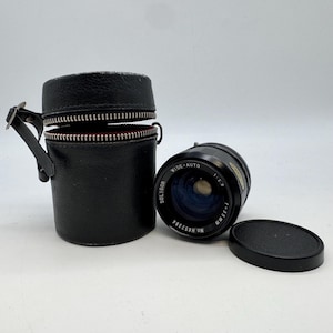 May include: A black Soligor Wide-Auto 1:2.8 f=35mm camera lens with a black lens cap and a black leather case. The lens case has a zipper closure and a small handle. The lens is marked with the text "WIDE-AUTO" and "SOLIGOR".