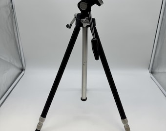 May include: A black and silver tripod with three legs. The tripod has a black head with adjustment knobs and a mounting plate. The legs are black with silver accents and adjustable feet. The centre column is silver.