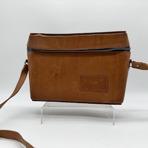 May include: A brown leather camera bag with a black zipper closure and a shoulder strap. The bag has a rectangular shape with a small, rectangular patch on the front. The leather shows signs of wear.