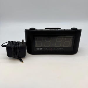 May include: Black digital alarm clock with a large display. The clock has a rectangular shape with rounded edges and the brand name "COBY" is visible. An AC adapter is included. The clock is designed for bedside or desk use.