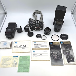 Puede incluir: Un conjunto de equipos de cámara Minolta Maxxum, que incluye un cuerpo de cámara negro, lentes, flash y manuales. La cámara tiene una correa gris y blanca. También hay varias tapas de lentes y filtros.