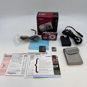 May include: A Casio Exilim digital camera in a maroon box with a battery, memory card, charger, power cable, AV cable, grey carrying case, and several printed manuals and accessory registration documents included in the set.