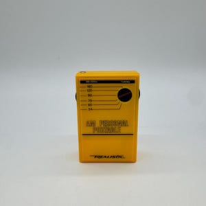 May include: A bright yellow rectangular AM personal portable radio with a black tuning dial and frequency scale printed on the front. The text AM PERSONAL PORTABLE and the brand name REALISTIC are clearly visible in black lettering on the front.