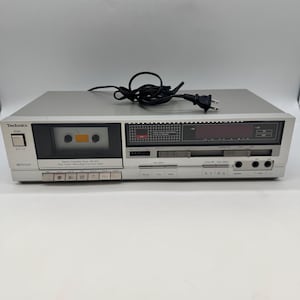 Puede incluir: Plataforma de casete estéreo Technics plateada con una cinta de casete insertada. La plataforma tiene una pantalla digital, varios botones y un cable de alimentación. El texto "Technics" es visible en la parte frontal.