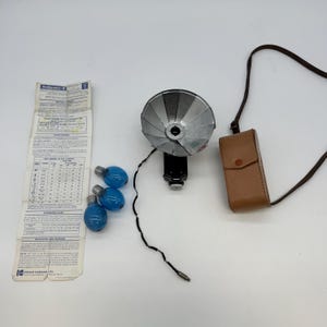 Puede incluir: Flash Kodak vintage con reflector plateado, base negra y estuche de cuero marrón. Incluye tres bombillas azules y una hoja de instrucciones Kodakcolor-II. El flash tiene un cable y un conector.