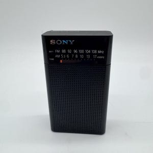 May include: A black Sony portable radio featuring a rectangular shape with a perforated speaker grille on the lower half. The upper section displays a frequency dial with FM and AM bands, a battery indicator, and a red tuning needle indicator.