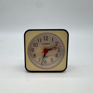 May include: A square, beige quartz alarm clock with a black border. The clock face has black numerals and hands, with a red second hand. The brand name "QUEMEX" is printed above the center.