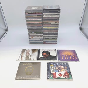 Op de afbeelding: Een verzameling muziek-cd's met twee hoge stapels doosjes en vijf individuele albums die ervoor zijn uitgestald. De albums bevatten titels van Tom Jones, Pavarotti en een compilatie getiteld O Canada! met stripfiguren.