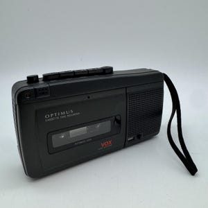 May include: A black Optimus cassette tape recorder featuring a built-in speaker, a top-mounted control panel with playback buttons, a clear cassette compartment window, and a black wrist strap. The device is labelled with Optimus and VOX text.