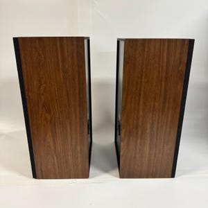 May include: Two vintage speakers with a wood grain finish. The speakers have a rectangular shape with black accents on the sides. The wood grain has a warm brown tone, and the speakers are positioned side by side.