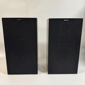 May include: Two black Sony speakers. Each speaker is a rectangular box with a circular speaker visible on the front. The word "SONY" is printed in white on the top of each speaker.