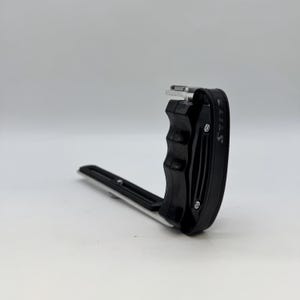 May include: Black camera grip with a silver base. The grip has a curved handle and a black strap with white text. The camera accessory is designed to provide a secure hold for photography.
