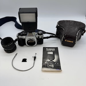 May include: A Canon AE-1 camera kit with a flash, lens, and case. The camera is silver and black with the Canon logo. The kit includes a black lens, a flash unit, a camera strap, a case, and a manual.