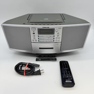 Könnte beinhalten: Ein silber-grauer, tragbarer Sony CD- und Kassetten-Player mit CD-Fach an der Oberseite, zentralem Digitaldisplay, zwei Lautsprechern und einem Kassettenfach an der Vorderseite. Enthält ein schwarzes Netzkabel und eine schwarze Sony-Fernbedienung.