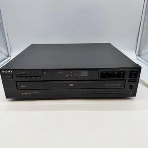 Puede incluir: Reproductor de CD Sony negro con cambiador de 5 discos. La unidad tiene una pantalla digital y varios botones de control. El número de modelo es CDP-C245. El panel frontal tiene el logotipo de Sony y las palabras "NEW HIGH DENSITY LINEAR CONVERTER".