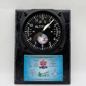 May include: A black and white altitude gauge clock on a black marble base. The clock face shows altitude in feet, with a plane logo. A blue plaque below commemorates a retirement, with text in English.