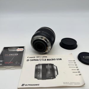 May include: A Canon EF-S 60mm f/2.8 Macro USM lens with two black lens caps, a DG filter, and a manual. The lens is black and silver with a ribbed design. The manual has a picture of the lens.
