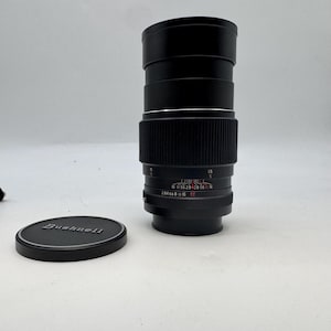 May include: A black camera lens with a ribbed focus ring and printed aperture markings in white and red. The lens features a depth of field scale and a separate black lens cap with the brand name Bushnell embossed in silver lettering on the top.