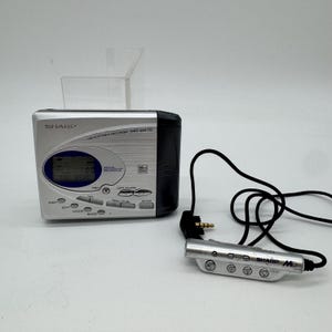 Puede incluir: Una grabadora de MiniDisc portátil Sharp MD-SR70 plateada con un panel lateral negro, una pantalla digital azul y varios botones de control. Incluye un mando a distancia con cable plateado a juego con cuatro botones circulares y un cable negro.