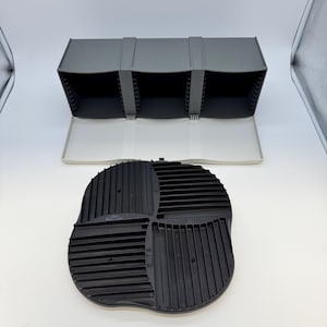 以下が含まれることがあります： 3つのオープンコンパートメントとヒンジ付きの蓋を備えたグレーのプラスチック製収納ユニット。収納容器内のアイテムを整理するための、平行な隆起を持つ4つの異なる象限を備えた、独立した黒い円形のインサートが付属しています。