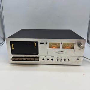 Puede incluir: Una pletina de casete estéreo TEAC A-100 plateada y negra con puerta de casete negra, dos vúmetros analógicos iluminados, un mando de nivel de grabación, varios botones, tomas de micrófono y auriculares e interruptores de palanca.