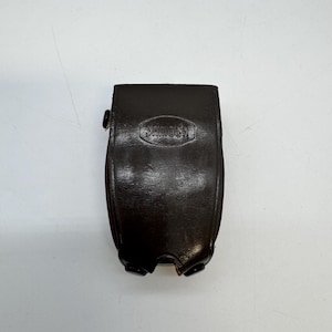 Könnte beinhalten: Eine dunkelbraune Ledertasche mit dem geprägten Wort "SAMOCH" auf der Vorderseite. Die Tasche hat eine gebogene Form und einen Schnappverschluss. Die Tasche scheint für eine Kamera oder ein anderes kleines elektronisches Gerät bestimmt zu sein.
