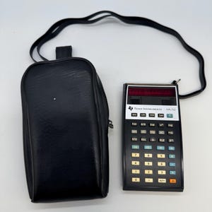Könnte beinhalten: Ein schwarzer Texas Instruments SR-50 wissenschaftlicher Taschenrechner mit einer schwarzen Ledertasche und einem schwarzen Riemen. Der Rechner hat ein rotes Display und verschiedene Funktionstasten.