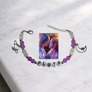 Peut inclure: Un bracelet de perles avec le mot "PLUTO" épelé en perles de lettres blanches. Le bracelet comporte des perles violettes, blanches et transparentes, avec un charm en forme de planète et un charm en forme de croissant de lune. Une petite affiche de film est au centre.