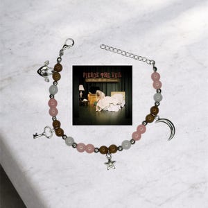 May include: A bracelet with pink, brown, and white beads, and silver charms including a key, a heart, a crescent moon, and a star. The bracelet is shown with a small album cover in the background.