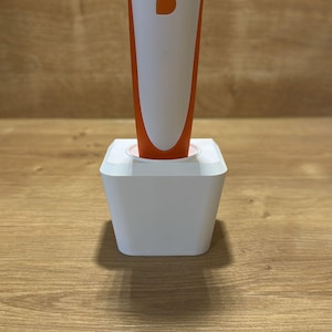Könnte beinhalten: Ein weiß-oranges Handgerät mit einer kreisförmigen Öffnung an der Oberseite und einem einzelnen orangefarbenen Knopf auf der Vorderseite, das senkrecht in einer weißen, quadratischen Ladestation steht. Das Gerät hat einen glatten, zylindrischen Körper.
