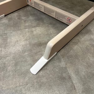 Könnte beinhalten: Eine helle Holzrahmenkonstruktion mit einer horizontalen schwarzen Metallstange und weißen Kunststofffüßen an der Basis. Ein rechteckiger Stützbalken zeigt aufgedruckte Sicherheitshinweise und ein kreisförmiges Symbol mit einer gehenden Person.