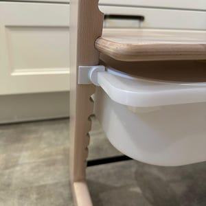 Könnte beinhalten: Ein hellfarbiger Hochstuhl aus Holz mit einem weißen Kunststofftablett und einem Holz Sitz. Der Hochstuhl hat ein stabiles Gestell und ist für ein Kind konzipiert. Das Tablett ist mit weißen Kunststoffclips an dem Stuhl befestigt.