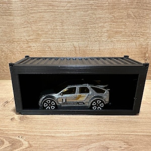 Könnte beinhalten: Ein silbernes Spielzeugauto mit gelben und schwarzen Akzenten wird in einem schwarzen rechteckigen Container ausgestellt. Das Auto hat die Nummer 5. Der Container steht auf einer Holzoberfläche.