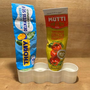 Könnte beinhalten: Zwei Tuben mit Gewürzen, eine blaue und eine gelbe, stehen in einem weißen Halter. Die blaue Tube ist mit "THOMY" und "MITTELSCHARFER DELIKATESS SENF" beschriftet. Die gelbe Tube ist mit "MUTTI" und "TOMATENMARK DREIFACH KONZENTRIERT" beschriftet.