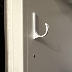 Könnte beinhalten: Ein weißer J-förmiger Haken, der an einer hellgrauen vertikalen Platte mit einer Reihe gleichmäßig verteilter kreisförmiger Löcher befestigt ist. Der Haken hat ein geschwungenes Design, das von der flachen Oberfläche absteht.