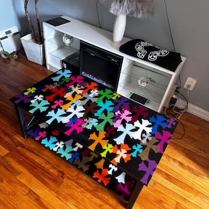 May include: A black coffee table with a lower shelf, decorated with a colorful cross pattern. The crosses are in various colors, including red, orange, blue, and purple. The table is made of wood and has a black frame.