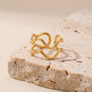 Könnte beinhalten: Ein goldfarbener Ring mit einem Doppelband-Design und einem durchgehenden, fließenden Wellenmuster. Der Ring hat eine polierte, metallische Oberfläche und eine offene Struktur, die zwei parallele, geschwungene Linien zeigt, die einen Lagen-Look erzeugen.