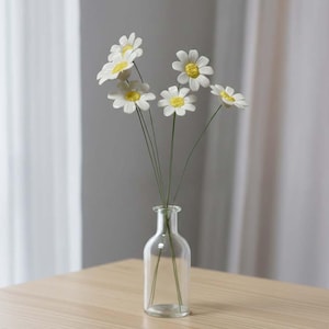 May include: A clear glass vase containing a small bunch of artificial daisies. The daisies have white petals and yellow centres. The vase is cylindrical with a narrow neck, and the flower stems are thin and green.