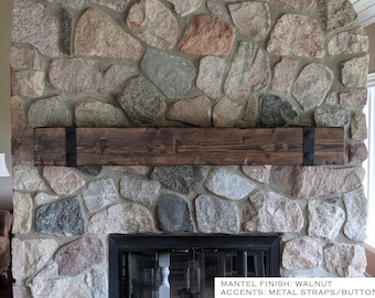 May include: A rectangular wooden mantel with a walnut finish, featuring two black metal straps on the left and right sides, each secured with two circular black buttons. The text on the image reads: MANTEL FINISH: WALNUT ACCENTS: METAL STRAPS/BUTTONS.