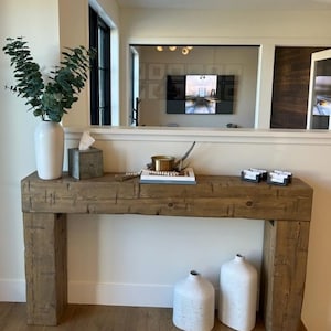 May include: A rectangular console table constructed from thick, weathered brown timber beams. The table features a flat top surface and two sturdy vertical legs. Two large white ceramic vases are placed on the floor beneath the table.
