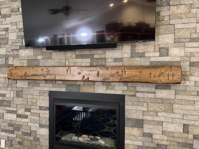 May include: A rectangular wooden fireplace mantel with a medium brown stain and a distressed, textured finish featuring visible wood grain, knots, and dark indentations, mounted horizontally on a wall covered in multi-tonal gray stone tiles.