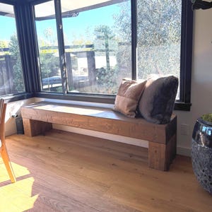May include: A long, rectangular wooden bench with a dark brown finish and thick, block-style legs. Two decorative throw pillows, one in a light tan shade and one in a dark charcoal gray, are placed on the top surface of the wooden bench.